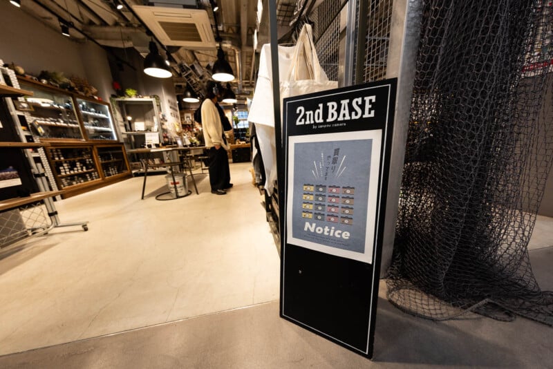 A black signboard reading "2nd BASE" with a notice is displayed at the entrance of a modern store, with shelves, products, and hanging lights visible inside. A person stands near the counter in the background.