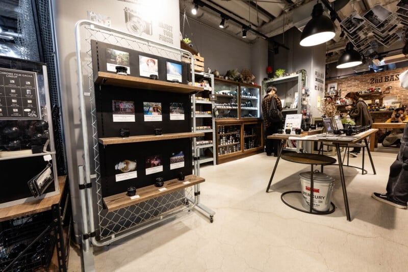A modern camera store interior with shelves displaying cameras and lenses, a table with a chair, and two people browsing. The store has bright lighting, industrial decor, and various photography equipment on display.