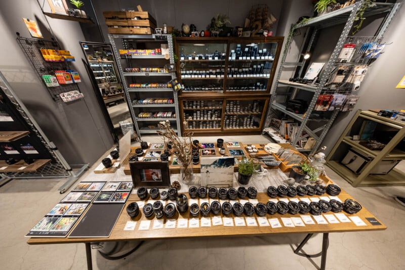 A display table in a camera shop with various camera lenses, film rolls, and accessories neatly arranged; shelves in the background are stocked with film, photographic gear, and art supplies.