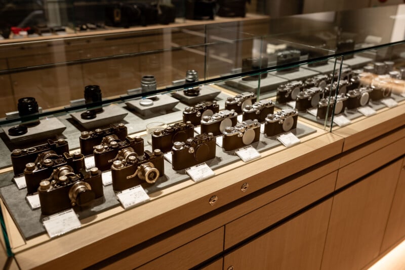 A display case with various vintage cameras and camera lenses arranged neatly on shelves, each with a small information card in front, inside a well-lit store or museum setting.