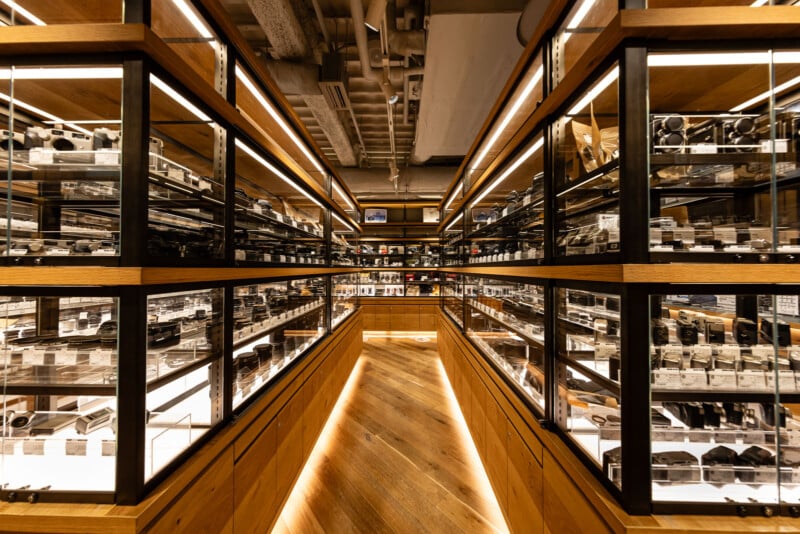 A well-lit shop aisle with glass display shelves on both sides, filled with assorted cameras and camera lenses. The wooden floor and ceiling pipes give the store a modern, industrial look.