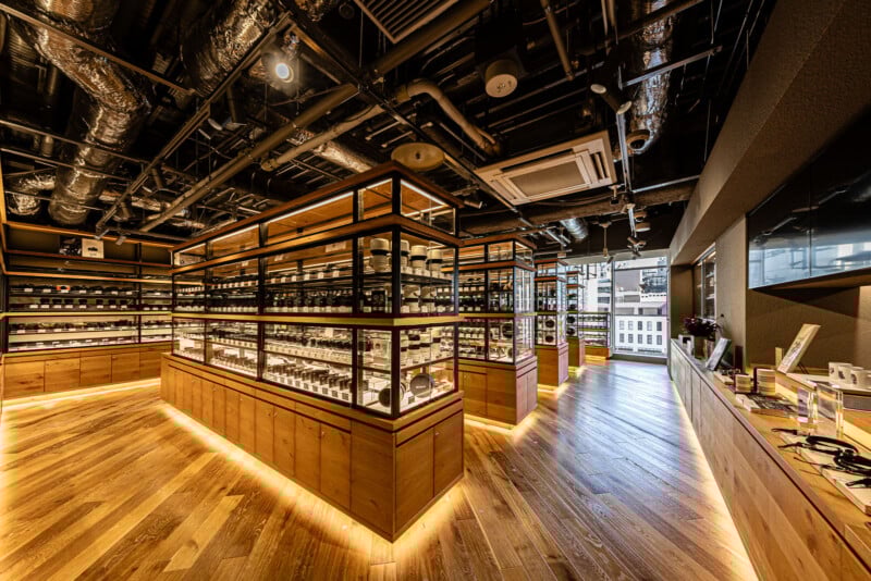 Modern camera store interior with wooden floors, glass display cases filled with camera lenses, and exposed industrial ceiling. Warm lighting highlights products and open space, with large windows at the back letting in natural light.