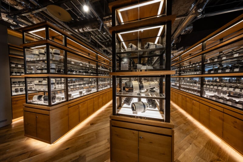 A well-lit room with wooden cabinets and glass display cases filled with numerous camera lenses and photography equipment, arranged neatly on shelves in a modern, organized setting.