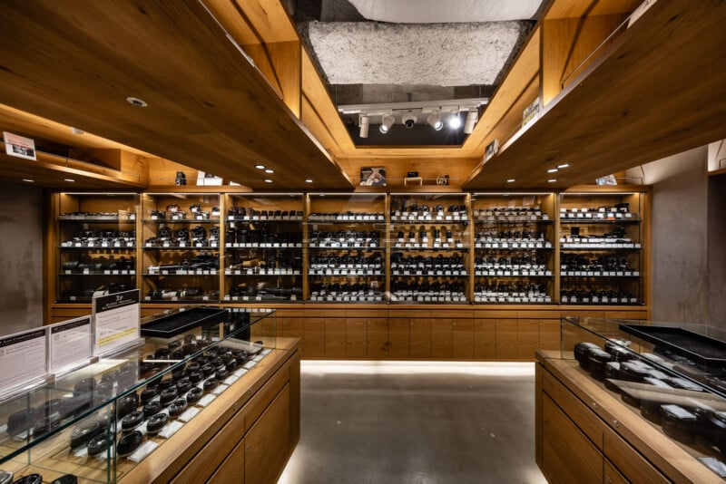 A camera store interior with wooden shelves and cabinets displaying many camera lenses and accessories, lit by ceiling spotlights. Glass display cases hold more equipment in the foreground.