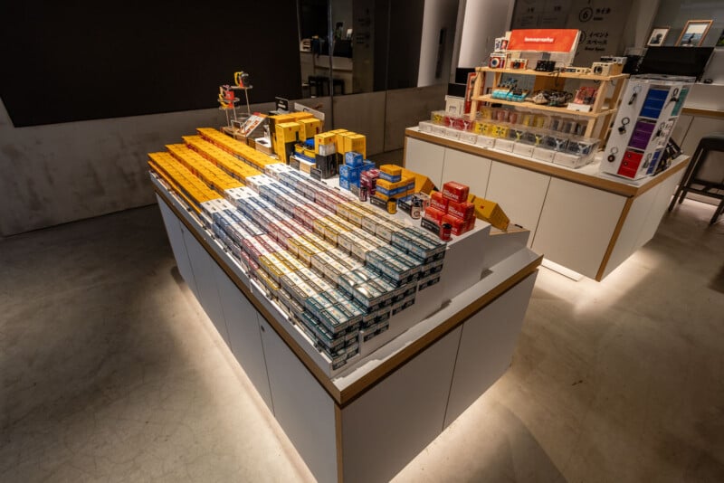 A well-lit store display features neatly arranged rows of colorful film boxes, including yellow, red, blue, and white packages, on a modern white counter with additional camera accessories on shelves behind.