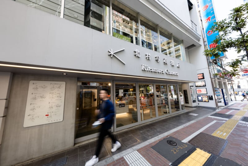A man walks past the entrance of Kitamura Camera, a modern camera store with large windows on a busy city street in Japan. The shop's name is displayed in Japanese and English on the building.