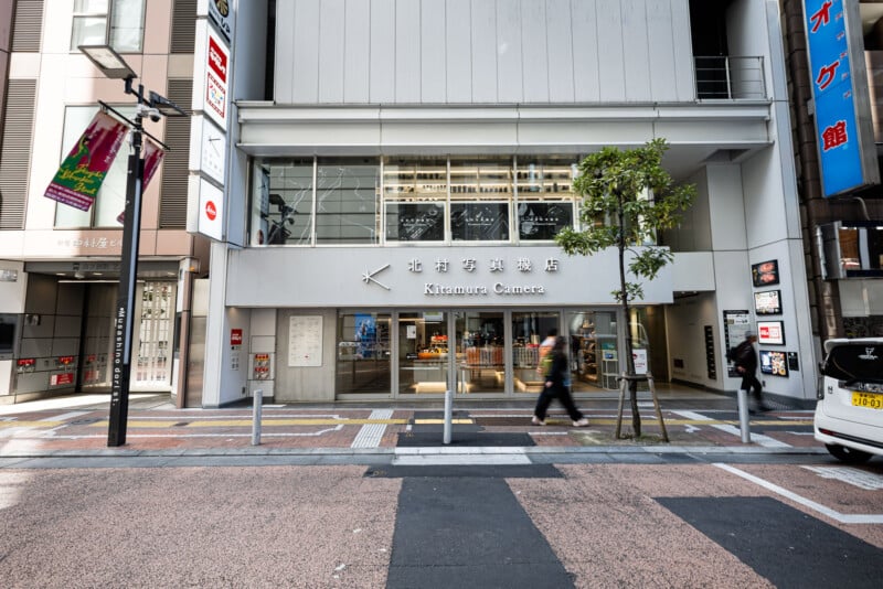 Street view of Kitamura Camera store in a modern building, with large windows, signs in Japanese, and people walking by on the sidewalk. There is a tree and street signs visible in front of the store.