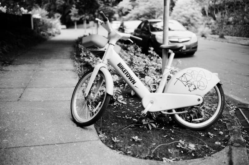 A Biketown rental bicycle with graffiti is locked to a pole on a sidewalk next to a street, with a parked car and trees in the background. The scene is in black and white.