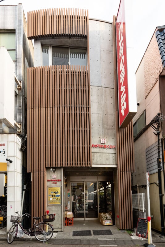 A modern two-story storefront in Japan with vertical wooden slats and a large red sign in Japanese above the entrance. Bicycles are parked outside, and the opened doors reveal shelves of goods inside.