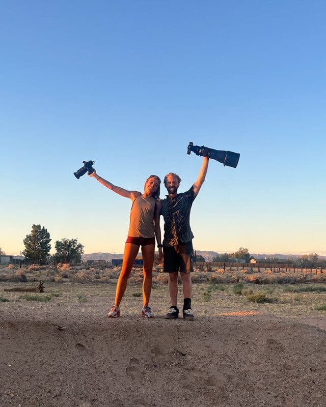 Two people stand side by side outdoors at sunset, each holding a camera with a large lens raised in one hand, smiling and posing in a desert-like landscape with clear blue sky.