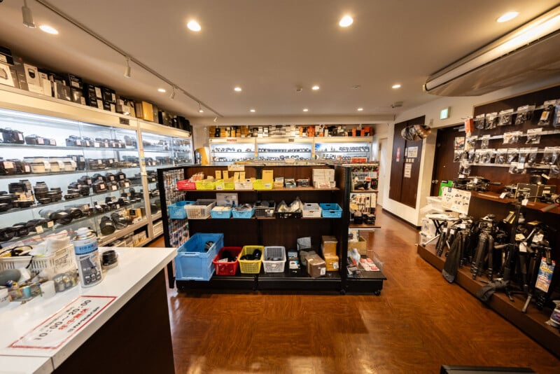 A well-lit camera store interior with shelves displaying cameras, lenses, photography equipment, and accessories. Bins of items are on a central table, and the floor is wooden.