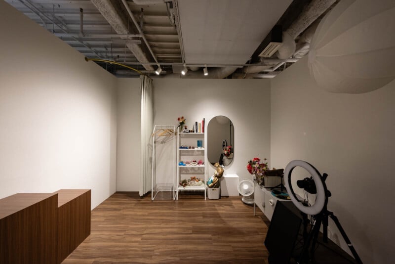 A photography studio with wooden floors, white walls, a ring light, camera, mirror, a clothing rack with hangers, shelves with props and flowers, and a fan, all under exposed ceiling pipes and spotlights.