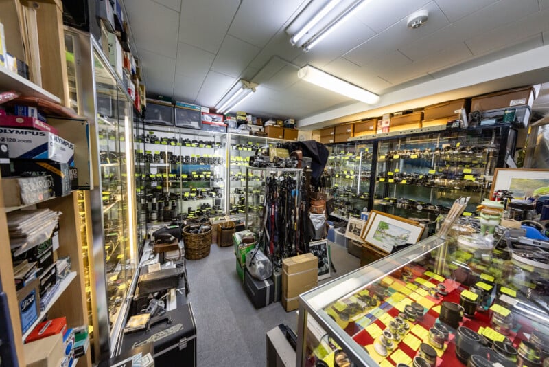 A cluttered camera shop interior with glass display cases filled with cameras, lenses, and accessories. Shelves line the walls, holding various camera equipment, boxes, and photography gear. Yellow price tags are visible throughout.