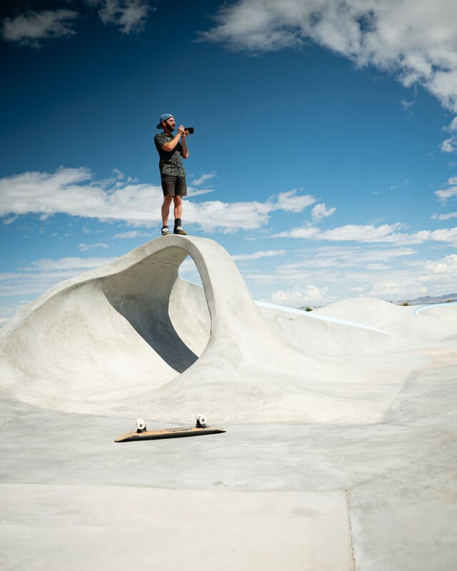 A man stands on top of a sculpted concrete ramp in a skatepark, holding a camera. A skateboard rests on the ground below him. The sky is bright blue with scattered clouds.