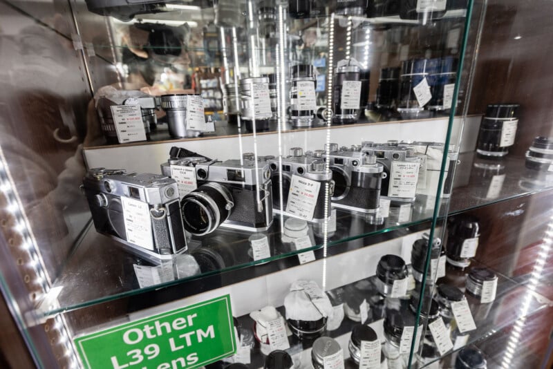 A glass display case with vintage film cameras and various camera lenses, each labeled with price tags. A green sign at the bottom reads "Other L39 LTM." The reflection shows a busy store interior.