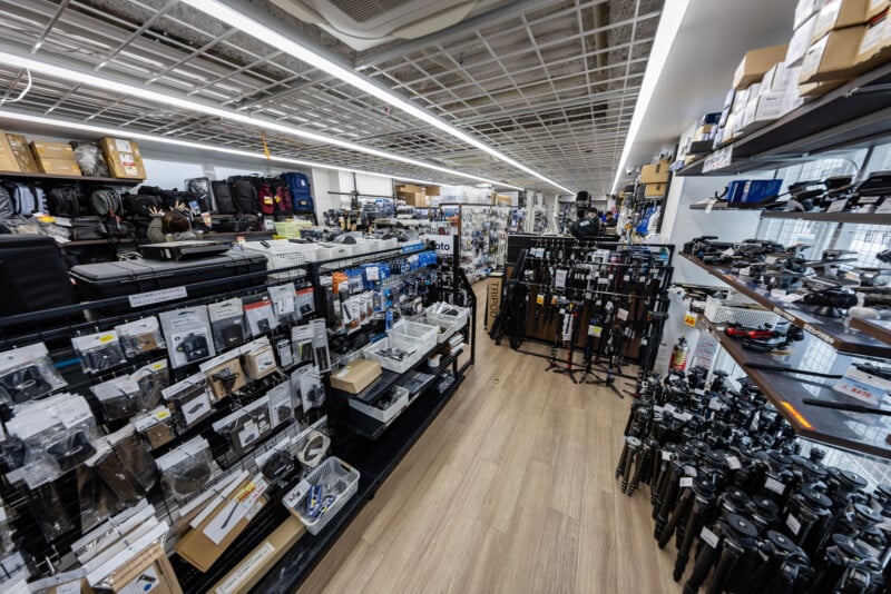 Wide-angle view of a well-lit camera store with shelves stocked with camera accessories, tripods, and equipment on both sides of a wooden-floored aisle. Boxes and gear are organized throughout the space.