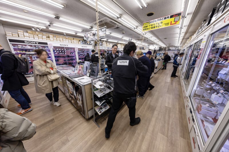 Customers and staff interact inside a brightly lit electronics store filled with display counters showcasing gadgets and camera gear. Signs in Japanese hang from the ceiling, and shelves are stocked with various products.