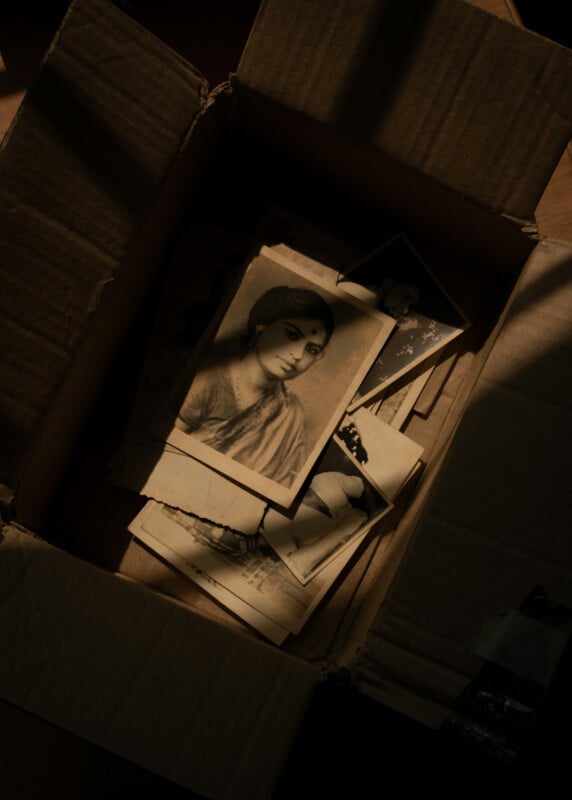 A cardboard box filled with old black-and-white photographs, with the top photo showing a woman in traditional attire, partially illuminated by soft light.