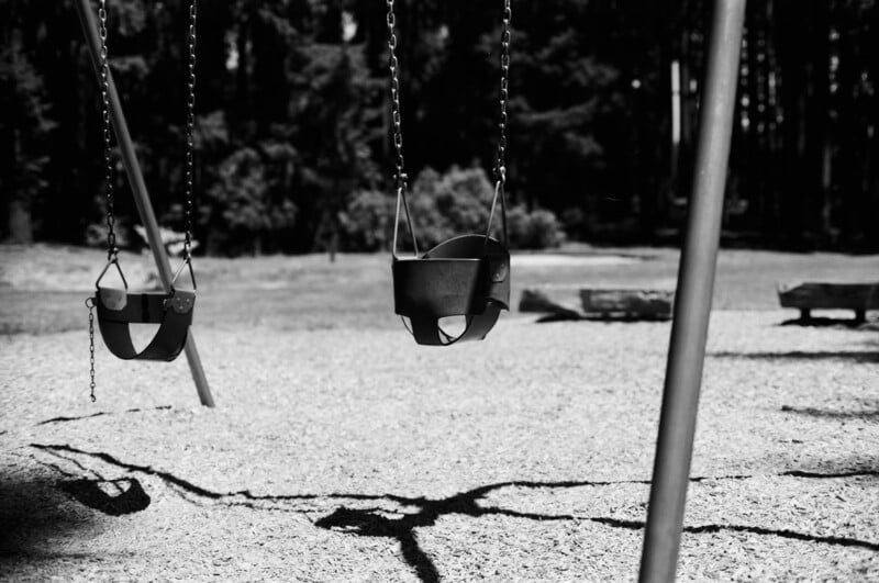 Black and white photo of two empty bucket swings hanging at a playground, casting distinct shadows on the gravel below, with trees and blurred park benches in the background.