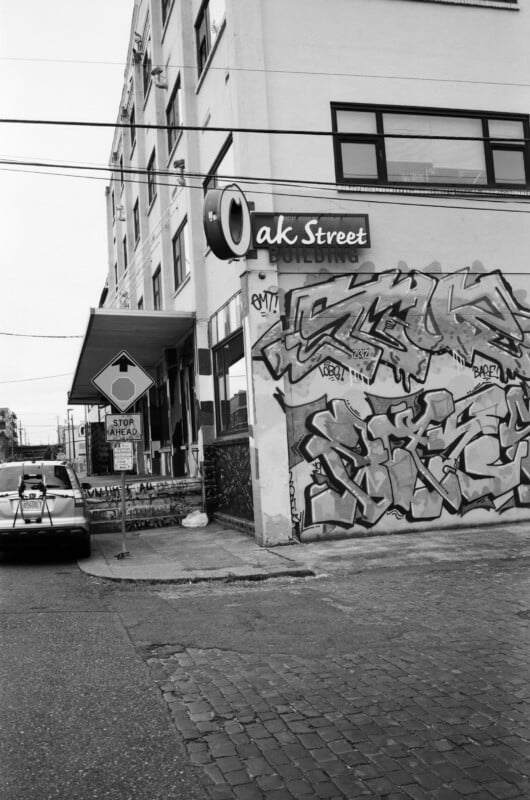 A city street corner with a building featuring a large "Oak Street" sign and colorful graffiti on its side. A taxi is stopped near a "Stop Ahead" road sign on a cobblestone street.