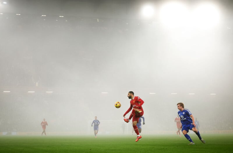 A soccer player in a red uniform jumps to control the ball mid-air, while a player in blue approaches. The field is covered in heavy fog, with stadium lights glowing in the background.