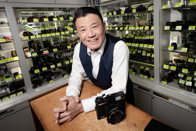A smiling man in a white shirt and navy vest stands behind a counter with a camera in front of him, surrounded by shelves filled with various cameras and photography equipment.