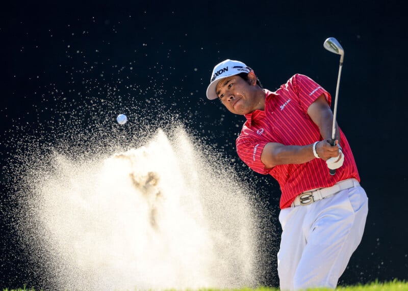 A golfer in a red shirt and white cap hits a shot from a sand bunker, sending sand and a golf ball into the air against a dark background.