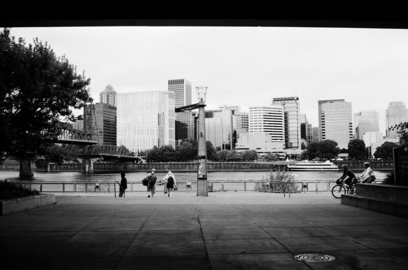 Black and white photo of people walking and biking along a riverside path, with a bridge and a city skyline of tall buildings in the background. Trees frame the left side of the image.