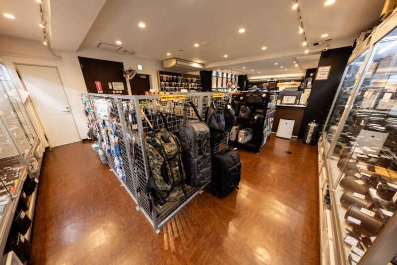 A well-lit store interior with shelves displaying various bags, backpacks, and outdoor gear. Glass display cases line the right wall, and the wooden floor adds warmth to the organized retail space.