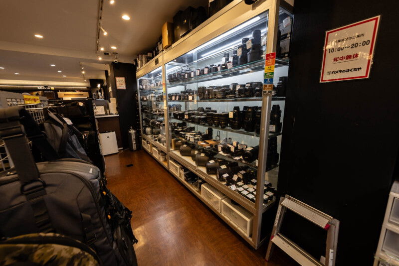 A glass display case filled with various camera lenses and equipment stands against a wall in a well-lit camera store, with backpacks and other gear visible on the left. A sign with Japanese text is mounted on the wall.