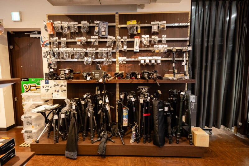 A store display with various camera accessories hanging on the wall and several tripods of different sizes standing on shelves below. The area is well-lit and organized.