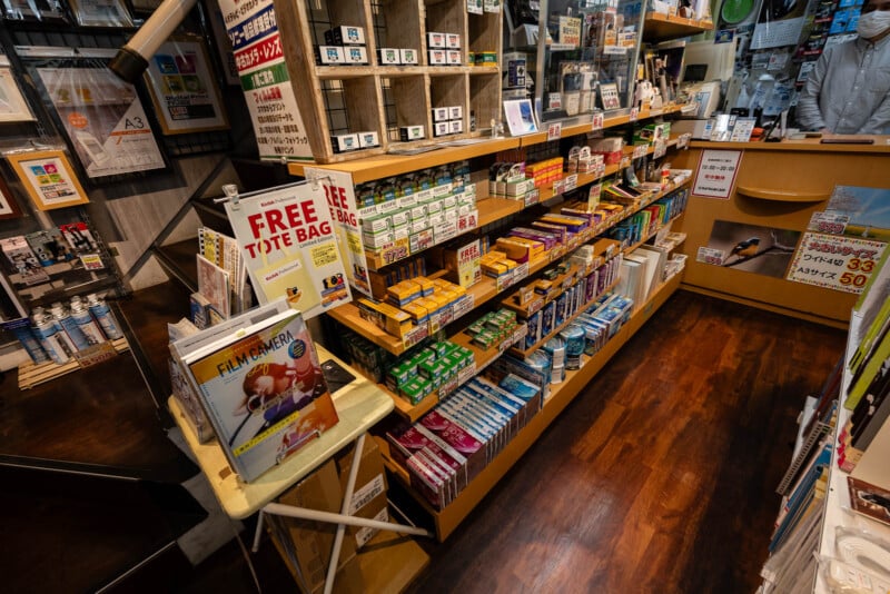 A display of camera film, batteries, and photography accessories in a well-lit shop with magazines, a “FREE TOTE BAG” sign, and a person behind the counter wearing a mask.