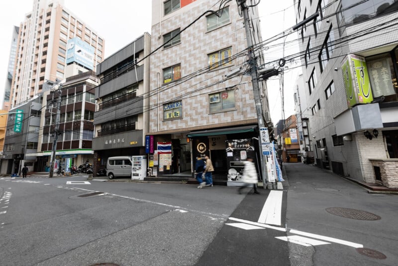 A street corner in an urban area with mid-rise buildings, a van parked on the side, overhead wires, and a few pedestrians walking near storefronts with Japanese signage.