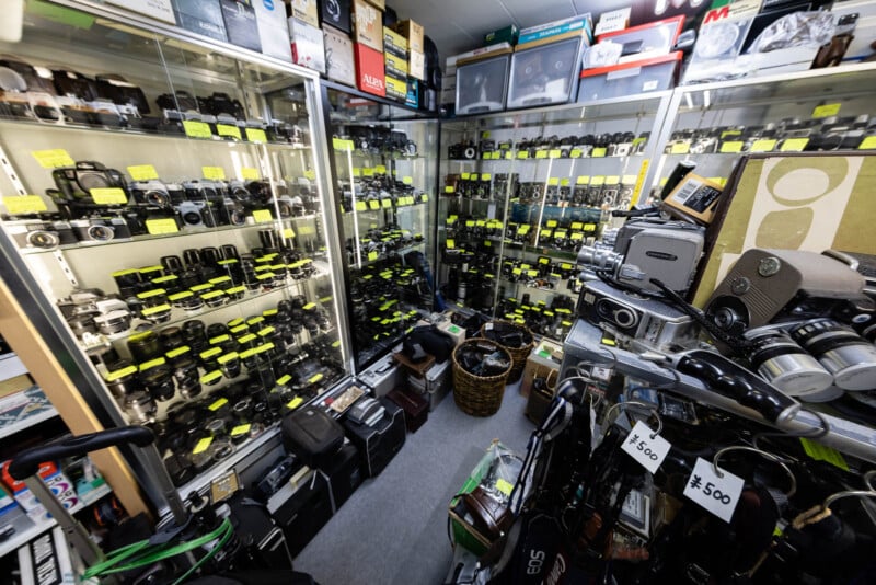 A small, crowded shop filled with vintage cameras, lenses, and camera equipment, neatly displayed on shelves and in glass cases. Many items have yellow price tags, and boxes and bags are stacked on the floor.