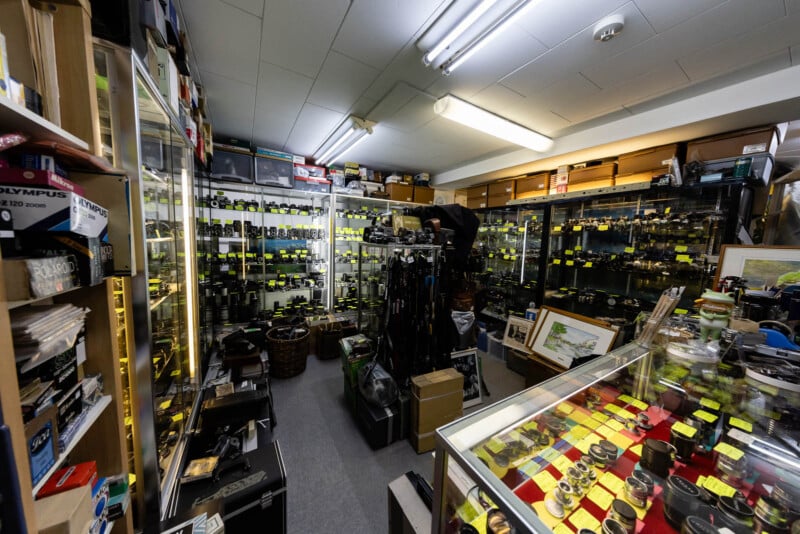 A cluttered camera shop filled with glass display cases showcasing cameras, lenses, and accessories, with shelves packed from floor to ceiling and yellow price tags visible on many items.