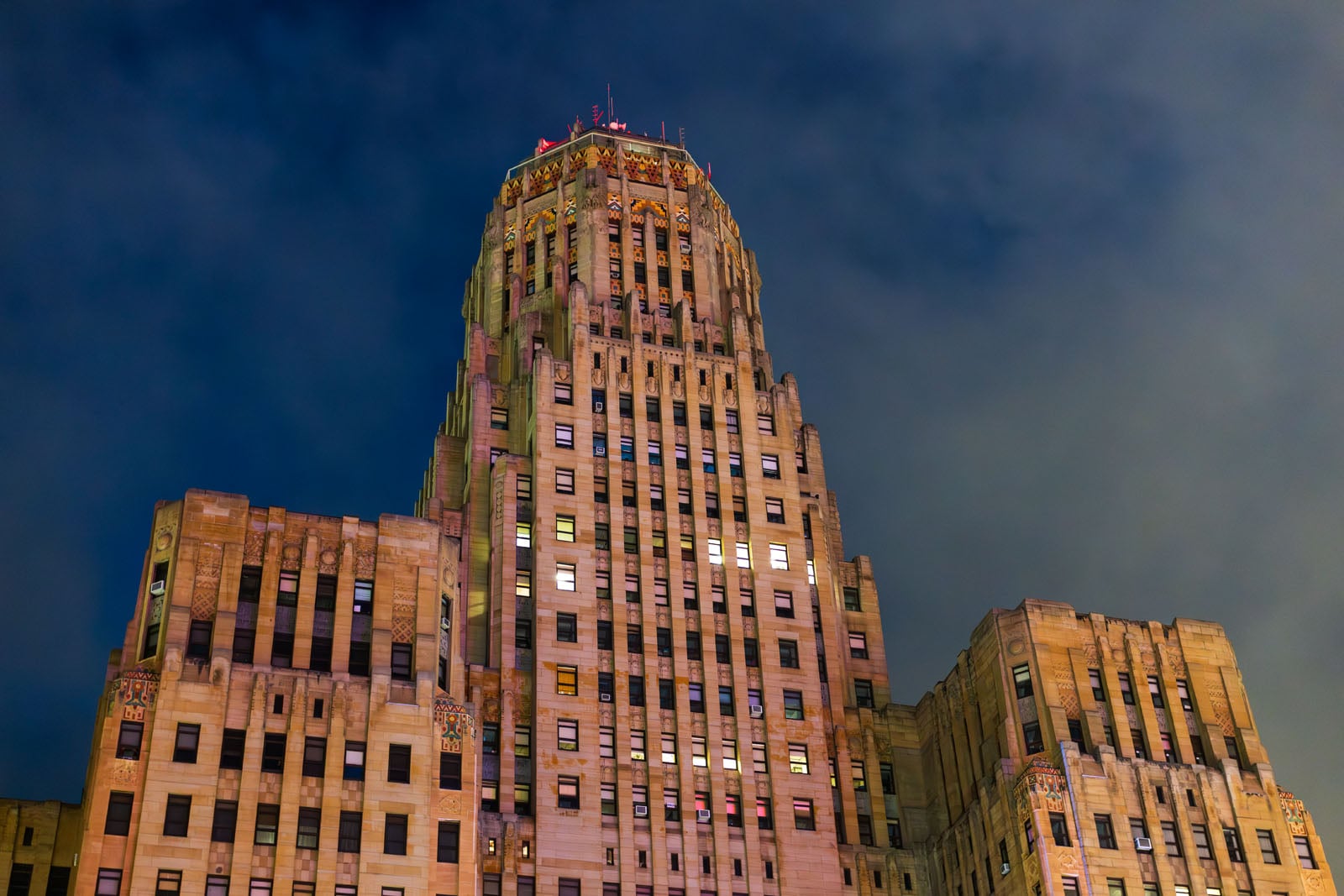Tall Art Deco skyscraper with intricate architectural details, warmly lit against a dark, cloudy night sky. The building features stepped layers and numerous illuminated windows.