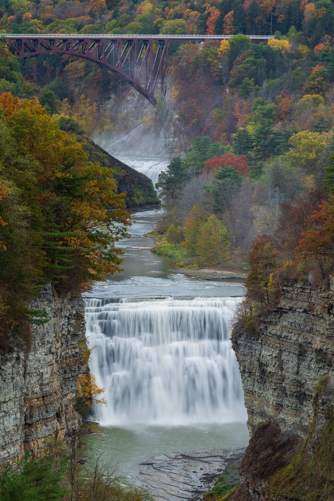 A river cascades over a wide waterfall in a rocky gorge surrounded by colorful autumn trees, with a red iron bridge spanning above the scene in the background.