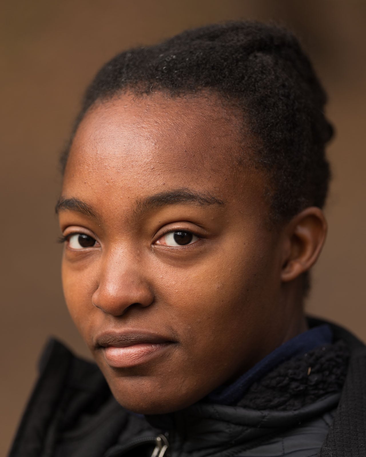 A person with short curly hair looks slightly to the side, wearing a dark jacket. The background is softly blurred, drawing attention to their calm expression.