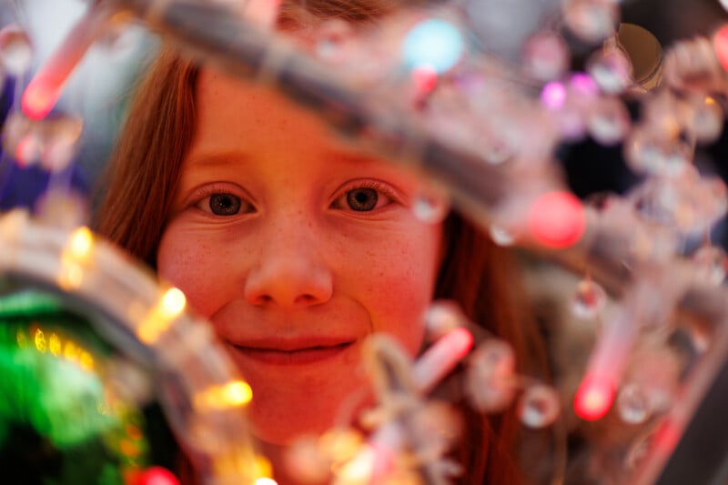 A young girl with long red hair and freckles is smiling, surrounded by colorful, glowing holiday lights and decorations, creating a festive and joyful atmosphere.