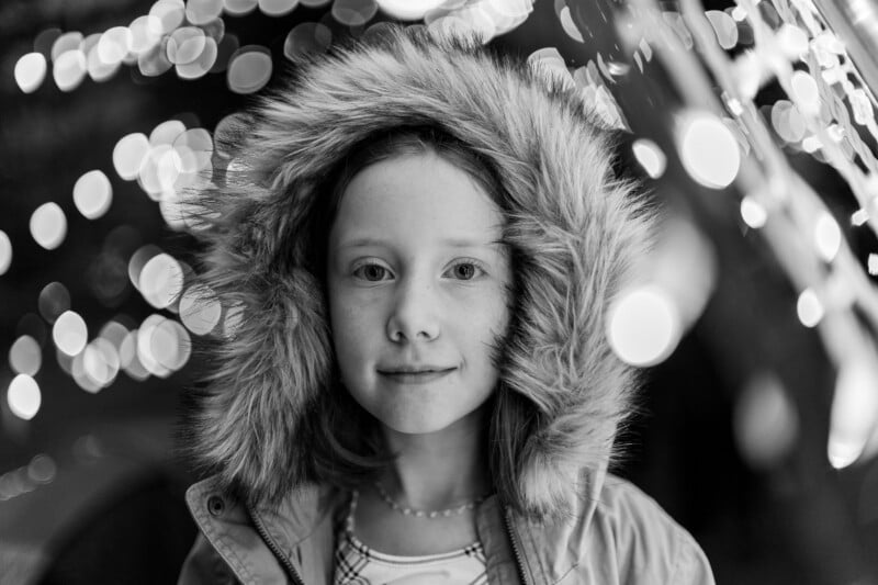 A young girl in a fur-lined hooded coat stands outdoors at night, surrounded by blurred festive lights. She looks directly at the camera with a calm expression. The image is in black and white.