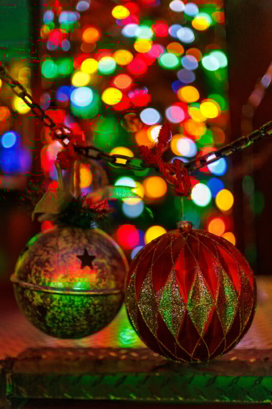 Two glittery Christmas ornaments, one silver and one red, hang from black chains with colorful, out-of-focus holiday lights creating a festive background.