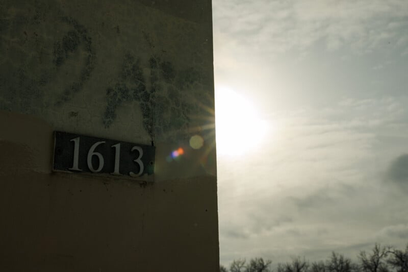 A weathered building corner displays the number 1613 as sunlight creates a lens flare, with a hazy sky and bare trees visible in the background.