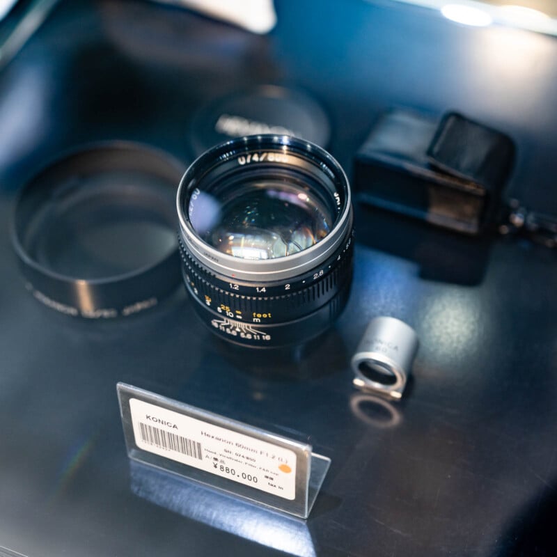 A Konica Hexanon 85mm f/1.2 camera lens is displayed on a glass surface with its cap, a small viewfinder, and a black pouch nearby. A product label with pricing is visible in front of the lens.