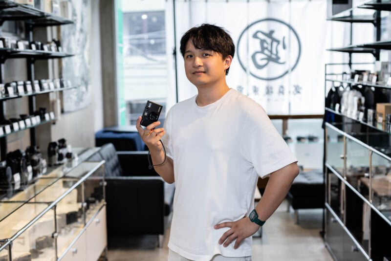A man in a white t-shirt stands in a modern store holding a small black product, surrounded by shelves displaying various items. The store has glass cases and a logo on a curtain in the background.