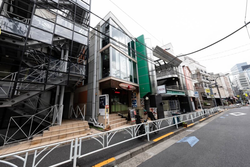 A street view of modern and narrow buildings in Tokyo, Japan, with signs, stairs, and café entrances lining the sidewalk beside a metal fence on a quiet, overcast day.