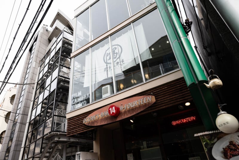 A modern, multi-story building with large glass windows. A wooden sign above the entrance reads "14 FOURTEEN" in English and Japanese. Reflections and power lines are visible on the windows.