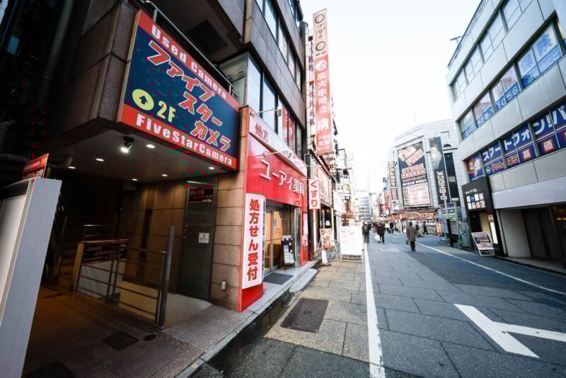 A city street in Japan lined with buildings featuring bright red and black signs in Japanese, including “Five Star Camera,” with a few pedestrians walking in the distance. The scene appears lively and urban.