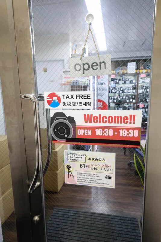 A glass door with "open" sign, "Tax Free" label, a "Welcome!!" sign showing open hours 10:30-19:30, and informational notices in Japanese and English, leading into a well-lit store.