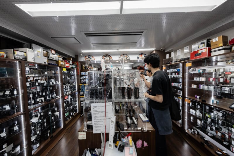 A man browses cameras and photography equipment in a well-lit shop filled with glass display cases and shelves packed with various cameras, lenses, and accessories.