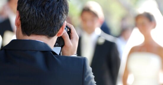 A photographer in a suit takes a photo of a bride and groom during an outdoor wedding ceremony. The couple, dressed in formal attire, are blurred in the background, while the photographer is in focus.